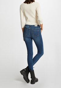 Dunkelblaue Skinny Jeans aus Denim mit glatter Struktur, feinen Nähten und einem Etikett am hinteren Bund. Kombiniert mit einem cremefarbenen Oberteil.