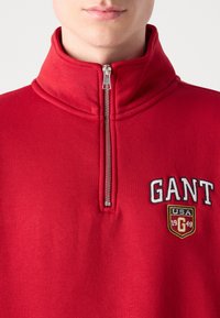 Pulover roșu cu guler înalt și închidere cu fermoar pe jumătate. Prezintă logo-ul "GANT" și o insignă cu „USA 1949” în culori contrastante și broderie.