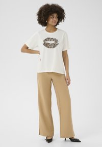 T-shirt crème avec un graphique de lèvres en imprimé léopard, associé à un pantalon large beige et des chaussures à talons noires. Tissu lisse, coupe décontractée.