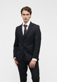 Fato preto ajustado com dois botões, bolso no peito, dois bolsos laterais, combinado com uma camisa branca e uma gravata roxa escura com bolinhas.