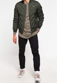 Vadderad, mörkgrön bomberjacka med ribbat krage och ärmslut. Bärs över en ljusgrön skjorta, tillsammans med svarta byxor och beige sneakers.