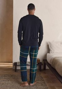 Pitkähihainen musta paita yhdistettynä vihreisiin ruutuhousuihin, joissa valkoisia ja keltaisia yksityiskohtia, esitetty takaa käsin paljain jaloin.