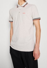 Polo gris clair avec un col classique, trois boutons et des poignets à rayures contrastantes noires et corail. Logo "BOSS" brodé sur la poitrine.
