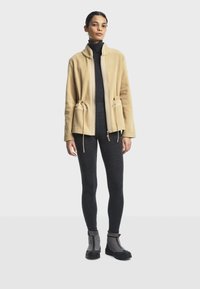 Veste en polaire beige avec un col haut, fermeture éclair à l'avant et cordon de taille. Associée à des leggings gris foncé et des bottes de pluie à la cheville.
