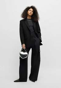 Blazer nero su misura, top nero aderente e pantaloni ampi abbinati a una borsa a forma di cuore argento. Tessuto liscio, design elegante e scarpe con punta.