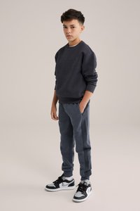 Tumši pelēks džemperis ar ģeometrisku tekstūru, apvienots ar atbilstošām sporta biksēm, kuru sānos ir kabatas. Melni un balti sporta apavi papildina tēlu.