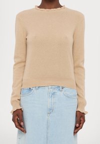 Pull beige à manches longues avec bords à volants, associé à une jupe en denim bleu clair, porté par une personne à la peau foncée.