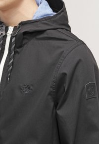 Homme portant une veste à capuche noire avec une fermeture éclair blanche et des cordons noirs, avec un petit logo circulaire sur la poitrine et un écusson sur la manche.