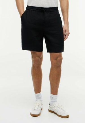 Mann trägt schwarze Shorts mit Kordelzug, weiße Sportsocken und weiße Turnschuhe, steht mit entspannt an den Seiten hängenden Händen.