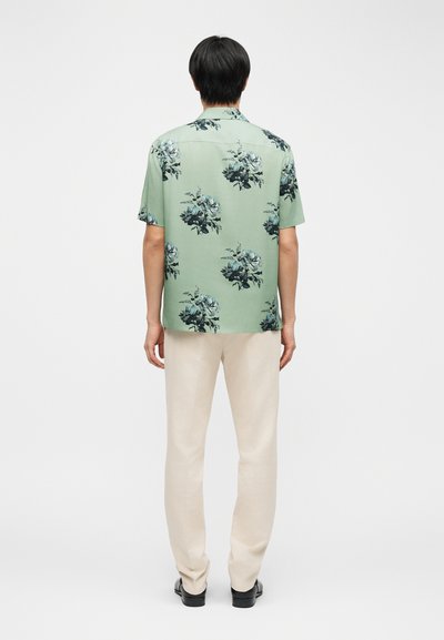 Άνδρας που φοράει ανοιχτό πράσινο κοντομάνικο πουκάμισο με σκούρο λουλουδάτο σχέδιο, μπεζ παντελόνι και μαύρα παπούτσια, στέκεται γυρισμένος με την πλάτη προς το λευκό φόντο.