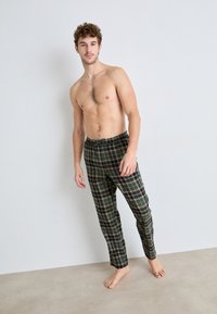 Gröna rutiga byxor med dragsko i midjan, med en mjuk bomullstextur och en strömlinjeformad passform, bärs av en modell med bar överkropp.