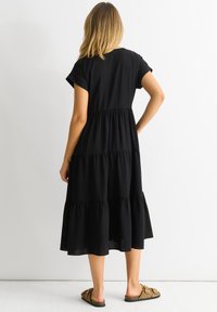 Black knee-length dress with short rolled sleeves, tiered design, elastic waistband, and flowing fabric. Worn with brown flat sandals.