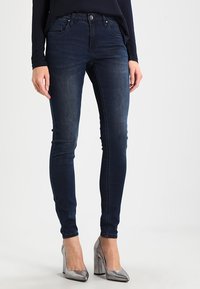 Skinny jeans in dark blue denim with a mid-rise waist, featuring five pockets, belt loops, and a smooth texture. Paired with silver high heels.