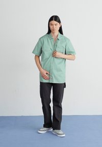 Cămașă scurtă verde deschis cu două buzunare la piept, asortată cu pantaloni în dungi întunecate și adidași gri. Design simplu, casual.
