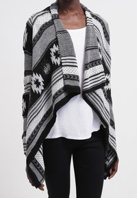 Černobílý vzorovaný kardigan s plášťovým límcem, otevřeným předním dílem a texturovaným pleteným vzorem. Látka volně splývá přes bílý top.