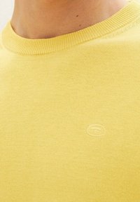 Gelber Pullover mit rundem Ausschnitt, gefertigt aus strukturiertem Material. Verfügt vorne über ein dezentes Logo, das Einfachheit im Design zeigt.