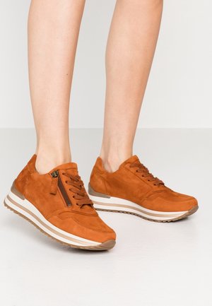 Sneakers in suede marrone con lacci e dettagli con zip laterale, indossati a piedi nudi su uno sfondo bianco.
