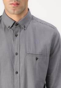 Gray checkered button-up shirt with a pointed collar, brown buttons, and a chest pocket featuring a horizontal stitch detail.