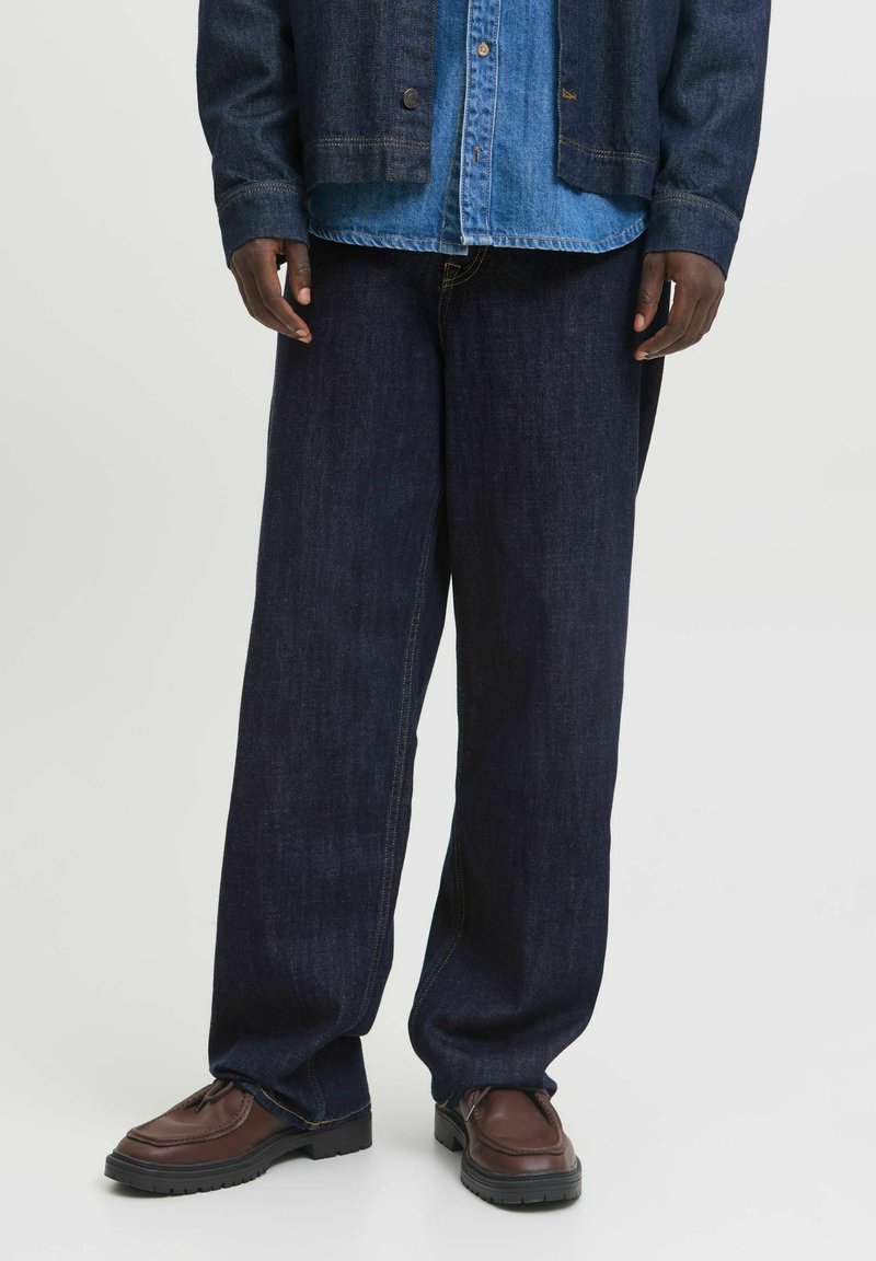 Pantalones de mezclilla azul oscuro con un diseño de pierna ancha, combinados con una camisa de mezclilla azul clara y mocasines marrones con suela negra.