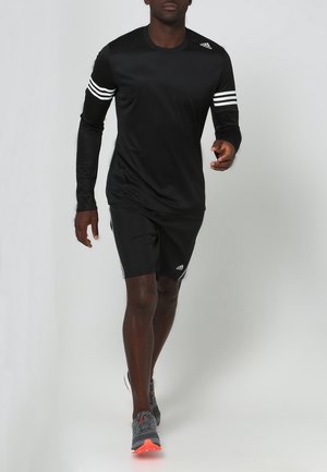 Musta pitkähihainen urheilupaita, jossa on valkoiset raidat hihoissa, yhdistettynä mustiin shortseihin ja harmaisiin juoksukenkiin, joissa on oransseja yksityiskohtia.