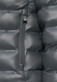Giacca gonfiabile di un grigio scuro con una texture liscia e lucida. Presenta una zip centrale con un tirante in tessuto e un motivo a quadri imbottito.