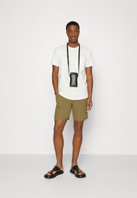 T-shirt gris clair, shorts vert olive et sandales noires. Une petite pochette noire pour téléphone pend d'une sangle noire autour du cou.