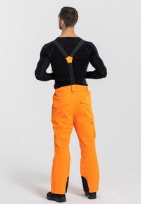 Oranje waterdichte broek met zwarte accenten, voorzien van verstelbare zwarte schouderbanden en twee achterzakken. Het materiaal lijkt duurzaam en stevig.