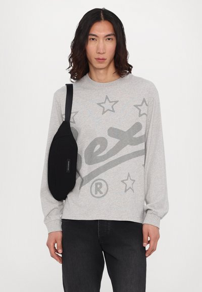 Gray long-sleeve shirt with star graphic, paired with black crossbody bag featuring a smooth texture and horizontal strap, worn over dark jeans.