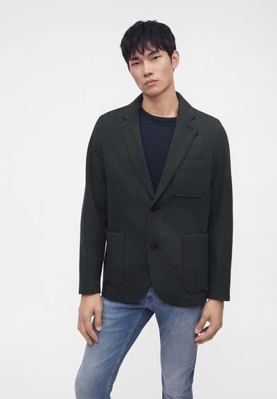 Jeune homme portant un blazer vert foncé sur une chemise noire et un jean bleu clair, debout sur un fond uni clair.