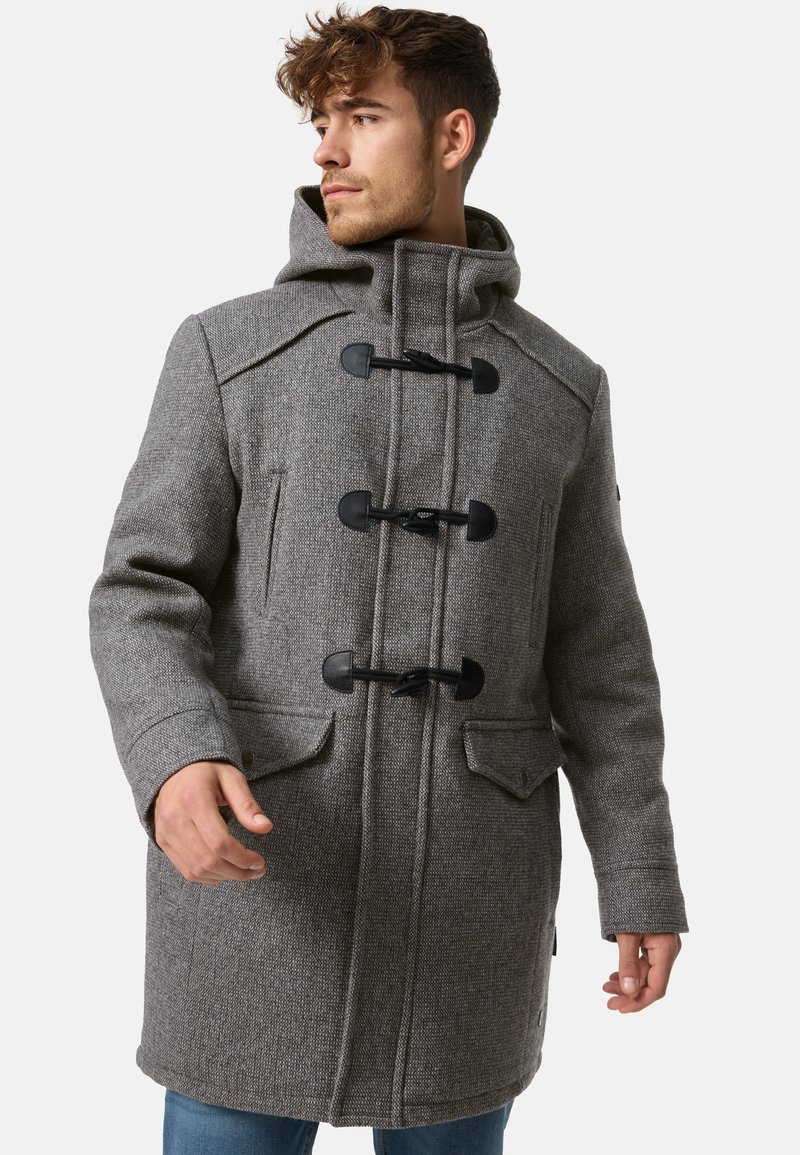 Homme portant un manteau duffle-coat gris à fermeture à brandebourgs avec capuche, debout et regardant sur le côté devant un fond uni.