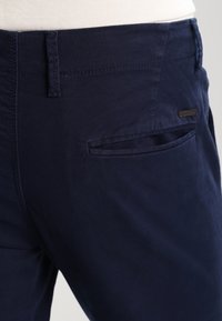 Pantaloni in cotone blu navy con fronte piatto, tasche laterali e un' etichetta del marchio discreta in vita, caratterizzati da una texture liscia ed uniforme.