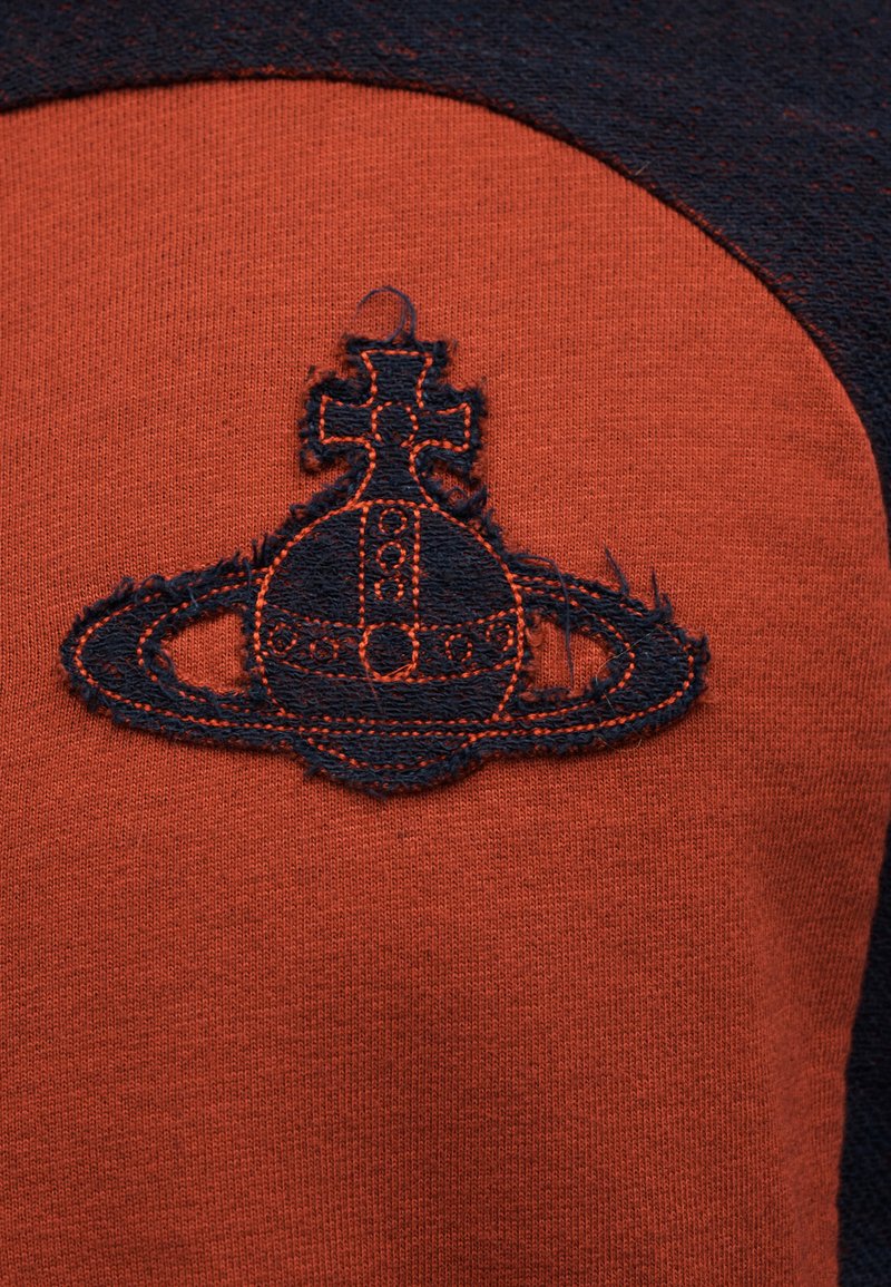Orange fabric with a dark navy embroidered orb and cross design featuring a ring around its base, resembling a stylized planet emblem.