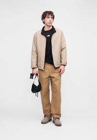 Μπεζ επενδυμένο μπουφάν με μαύρο γιακά, μαύρο μπλουζάκι με λογότυπο, καφέ παντελόνι και ανοιχτό γκρι μποτάκια. Κρατάει μια μικρή μαύρη τσάντα.