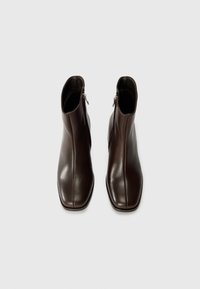 Bottines en cuir marron avec un design épuré, bout carré et fermetures à glissière sur les côtés. Texture lisse et lignes minimalistes sur un fond uni.