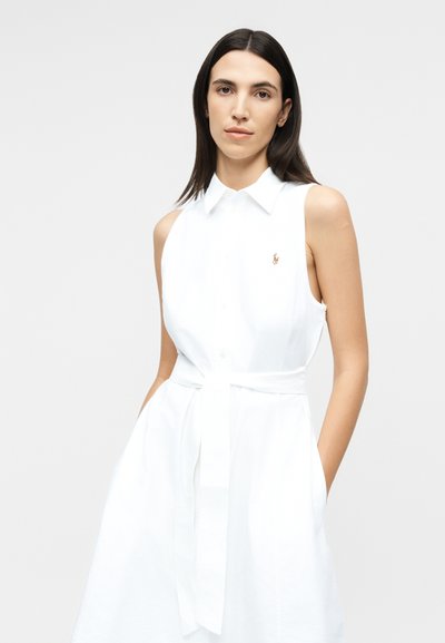 Sötét, egyenes hajú nő ujjatlan fehér, galléros ruhát visel derékszalaggal, és világos, egyszínű háttér előtt áll.