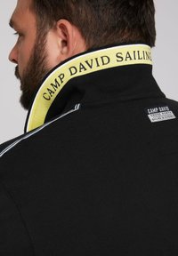 Black polo shirt with a high collar featuring a yellow "CAMP DAVID SAILING" band and a small brand label on the back, textured fabric.