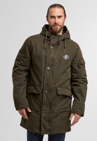 Olivgrüne Parkajacke mit Kapuze, großen Fronttaschen, Reißverschluss- und Knopfverschlüssen sowie einem Aufnäher an der linken Ärmel.