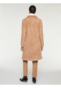 Cappotto in pelliccia sintetica color sabbia con colletto, maniche lunghe e taglio dritto, abbinato a pantaloni beige e stivali con tacco neri.