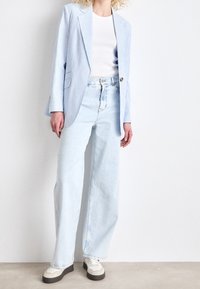 Person wearing light blue pinstripe blazer, white ribbed top, wide-leg light blue jeans, and beige sneakers standing against a white wall.