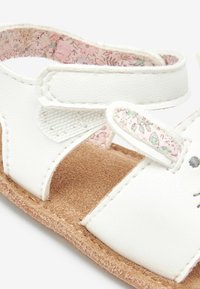 Sandales synthétiques blanches avec une surface texturée, ornées d'une doublure à motifs floraux et d'une semelle intérieure douce marron, fermées par une sangle auto-agrippante.