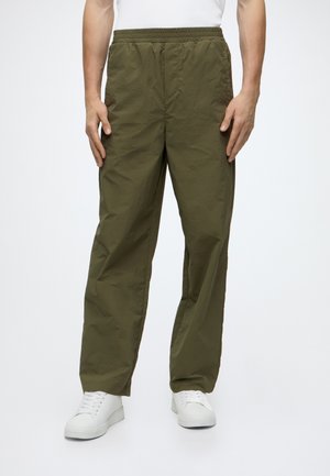 Homme portant un pantalon vert olive à taille élastique et des baskets blanches, debout les mains détendues le long du corps.