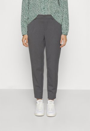 Grünes langärmeliges Hemd mit weißen Punkten, in graue, knöchellange Hose gesteckt, kombiniert mit weißen Turnschuhen an einer stehenden Person.