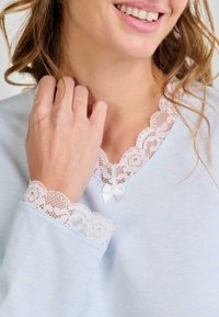 Primer plano de una mujer sonriendo, vistiendo una blusa de manga larga azul claro con detalles de encaje blanco y un pequeño lazo en el escote.