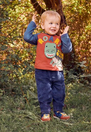 Ein Kind trägt ein rot-blaues Langarmshirt mit einem Dinosaurier-Motiv und dem Text "ROAR!", kombiniert mit einer marineblauen Cordhosen und bunten Sneakern.