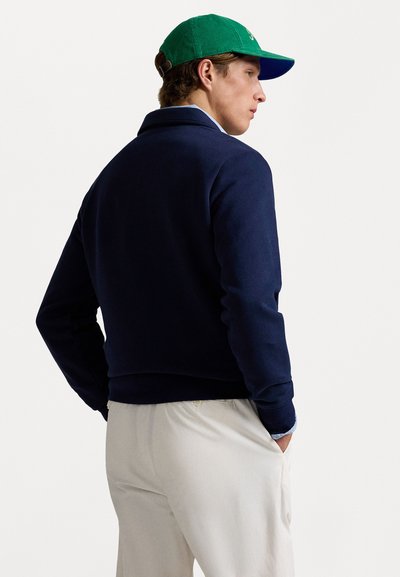 Sudadera azul marino con cuello alto y mangas largas, combinada con pantalones de color claro. Una gorra verde completa el conjunto.