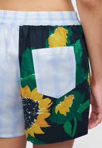 Άποψη από τη μέση έως το μέσο του μηρού ενός σορτς με ελαστική μέση, με μεγάλο μοτίβο ηλιοτροπίου και λευκή πίσω τσέπη με διακριτικό λογότυπο Moschino Jeans.