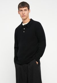 Schwarzer Strickpullover mit Polo-Kragen, drei Knöpfen, gerippten Bündchen und Saum, getragen über einem weißen Hemd, kombiniert mit karierten Hosen.