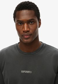 Sudadera de algodón gris oscuro con cuello redondo y logo bordado "SUPERDRY®" en el pecho. Textura suave y diseño casual.