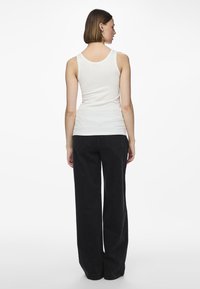 Woman with short brown hair wearing a white sleeveless top and black wide-leg jeans, standing facing away against a white background.