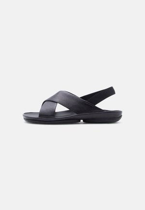 Sandalo nero in pelle con cinturini incrociati e cinturino posteriore elastico, suola piatta imbottita, vista laterale su sfondo bianco semplice.